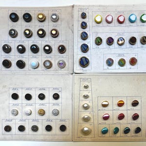 May include: Four rectangular display cards featuring an assortment of buttons in various sizes, shapes, and colors. The buttons are arranged in rows and columns with handwritten identification numbers printed on the off-white paper backing.
