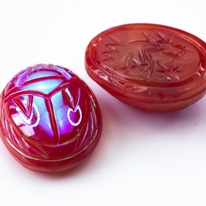May include: Two red glass scarab beetle charms with iridescent detailing. One charm is a flat oval with a carved design, the other is a three-dimensional scarab beetle.