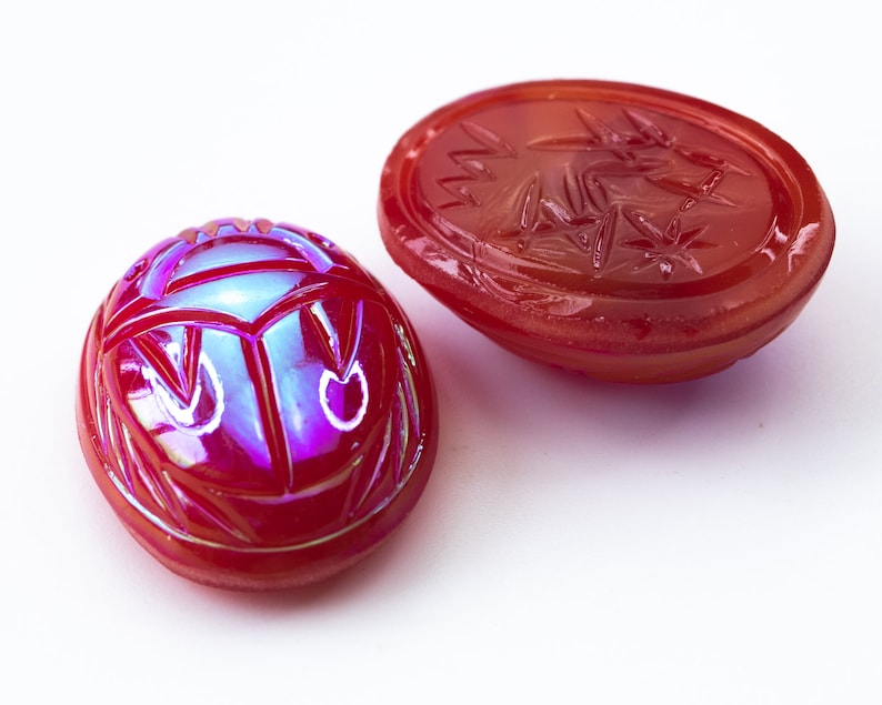 May include: Two red glass scarab beetle charms with iridescent detailing. One charm is a flat oval with a carved design, the other is a three-dimensional scarab beetle.