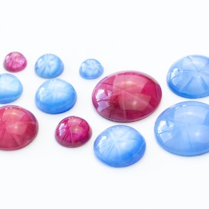 Pode incluir: Conjunto de pedras preciosas cabochão em forma de estrela em tons de azul e rosa. As pedras preciosas estão dispostas sobre uma superfície branca.