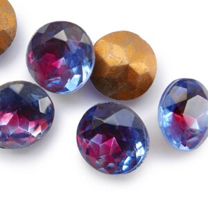 May include: Assortment of six faceted glass gems in shades of blue and purple. The gems are round and have a shiny, reflective surface.