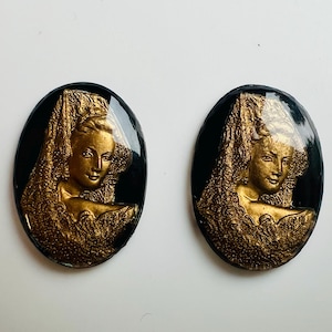 May include: A pair of oval earrings with a vintage design. Each earring has a gold-coloured relief of a woman's profile set against a black backdrop. The woman is wearing an elaborate headdress, with a classic, elegant look.
