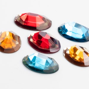 May include: Six oval-shaped rhinestones in various colors, including red, orange, and blue. The rhinestones are arranged in a semi-circle on a white background.