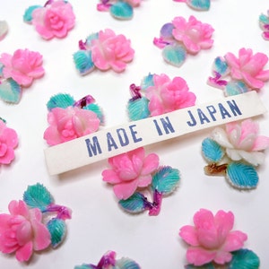 May include: A collection of pink and white plastic flower embellishments with blue and green leaves. The flowers are arranged on a white surface with a white label that reads "MADE IN JAPAN".