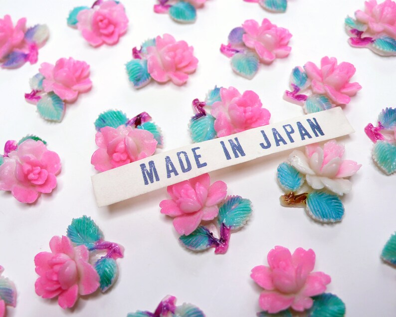 May include: A collection of pink and white plastic flower embellishments with blue and green leaves. The flowers are arranged on a white surface with a white label that reads "MADE IN JAPAN".