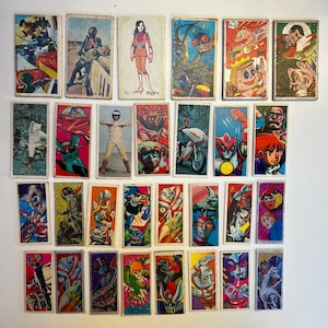 May include: A collection of vintage Japanese trading cards featuring colorful illustrations of anime characters, robots, and superheroes. The cards have a rectangular shape and are arranged on a white surface. The artwork includes various action poses and vibrant color schemes.