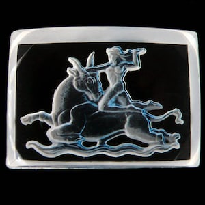 Puede incluir: Un pisapapeles de vidrio transparente con un diseño en relieve de una persona montando un toro. El diseño es de color azul claro.