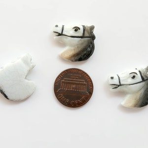 Puede incluir: Tres figuras de cer&aacute;mica con forma de cabeza de caballo en blanco, gris y negro, con una moneda como referencia de tama&ntilde;o. Las cabezas de caballo est&aacute;n detalladas con bridas y rasgos faciales. Una est&aacute; rota. Sobre fondo blanco.