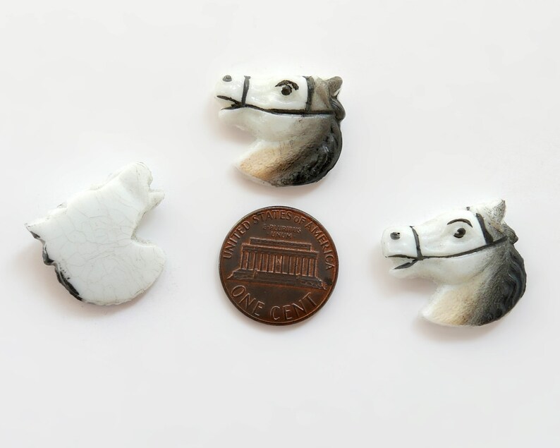 Puede incluir: Tres figuras de cer&aacute;mica con forma de cabeza de caballo en blanco, gris y negro, con una moneda como referencia de tama&ntilde;o. Las cabezas de caballo est&aacute;n detalladas con bridas y rasgos faciales. Una est&aacute; rota. Sobre fondo blanco.