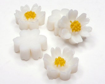 May include: Four white ceramic flower-shaped decorations with yellow centers. The flowers have five petals and are arranged on a white surface.
