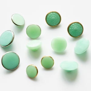 Puede incluir: Una colección de 12 botones de vidrio verde en varios estilos, incluyendo diseños redondos, planos y en relieve. Algunos botones tienen un borde plateado, mientras que otros tienen un borde dorado.