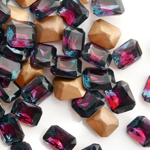 May include: A collection of dark purple and teal iridescent glass gems in a rectangular shape. The gems are scattered on a white surface.