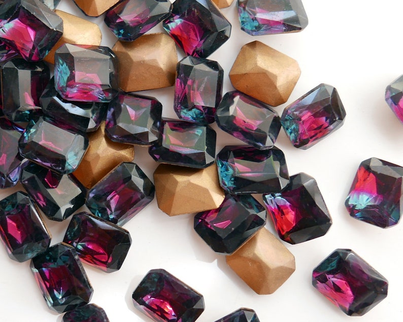 May include: A collection of dark purple and teal iridescent glass gems in a rectangular shape. The gems are scattered on a white surface.