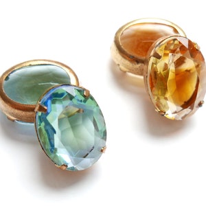 Puede incluir: Dos gemelos dorados de estilo vintage. Uno presenta una gran gema ovalada, facetada, azul-verdosa. El otro gemelo tiene una gema de color amarillo dorado. Los gemelos se muestran sobre un fondo blanco.