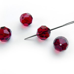 Op de afbeelding: Vier facetgeslepen, dieprode kralen met een naald die door één ervan is geregen. De kralen zijn gelijkmatig verdeeld op een witte achtergrond. De kralen hebben een geometrische vorm en zijn waarschijnlijk bedoeld voor het maken van sieraden of knutselen.