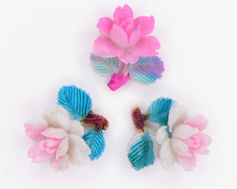 May include: Three decorative flower embellishments with pink and white petals and blue and green leaves. The flowers are made of a smooth, glossy material.