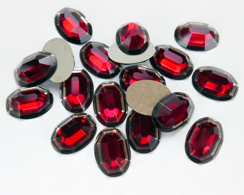 May include: A collection of red oval-shaped rhinestones with a faceted design. The rhinestones have a dark, almost black, backing.