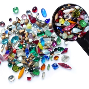 以下が含まれることがあります： 白い表面に、さまざまな色のガラス製のラインストーンとビーズが散らばっています。黒い計量スプーンに、さらにラインストーンとビーズが満たされ、山の上に傾けられています。