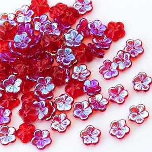 Op de afbeelding: Een grote stapel rode, iriserende, bloemvormige glazen kralen. De kralen hebben een glanzend, reflecterend oppervlak en zijn verspreid over een witte achtergrond.