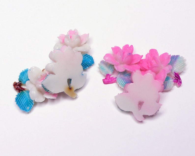 May include: Two sets of pink and white flower-shaped resin embellishments with blue and iridescent accents. The embellishments are perfect for adding a touch of whimsy to crafts and projects.