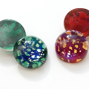 Op de afbeelding: Vier ronde glazen cabochons met een gespikkeld patroon. Eén is rood, één is groen en twee zijn iriserend met gouden vlokken.