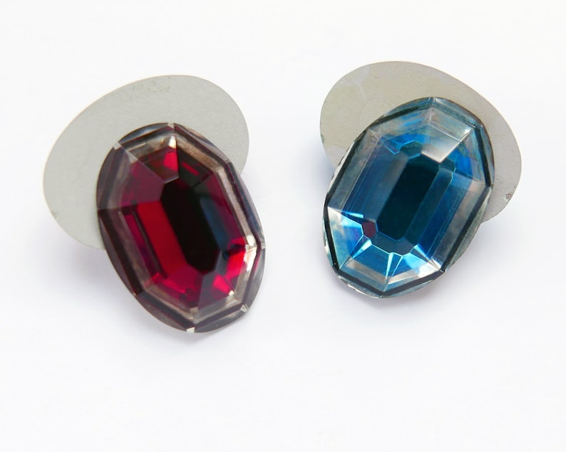 May include: Two oval-shaped glass rhinestones, one red and one blue, on white backgrounds.