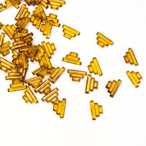 May include: A collection of small, amber-colored, rectangular beads scattered on a white surface. The beads have a layered, stepped design, resembling miniature architectural elements. Ideal for jewelry making or craft projects.