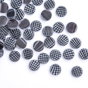 May include: A collection of small, round glass beads. The beads have a mix of patterns, including black and white checkered designs and swirling gray and black patterns. The beads are scattered on a white surface.