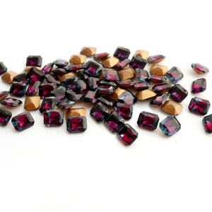 May include: A pile of dark purple and iridescent square-shaped rhinestones with gold settings.