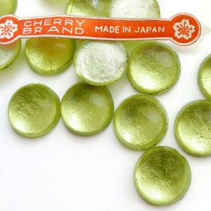 Puede incluir: Un grupo de 10 botones redondos de vidrio verde con un acabado metálico. Los botones están esparcidos sobre una superficie blanca. Una etiqueta dice "CHERRY BRAND MADE IN JAPAN".