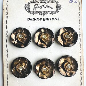 Peut inclure: Un ensemble de six boutons ronds sur une carte blanche marquée Fairy Queen Deluxe Buttons et Made in Czechoslovakia. Chaque bouton présente un motif de rose métallique sombre avec une pierre centrale claire, disposés en deux rangées de trois.