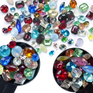 May include: Two black measuring spoons filled with colourful glass gems. The gems are in various shapes and sizes, including round, oval, and square. The gems are a mix of red, blue, green, purple, and clear.