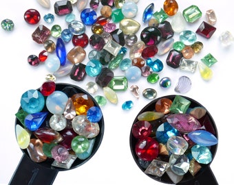 Puede incluir: Dos cucharas medidoras negras llenas de gemas de vidrio de colores. Las gemas son de varias formas y tamaños, incluyendo redondas, ovaladas y cuadradas. Las gemas son una mezcla de rojo, azul, verde, morado y transparente.