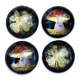 Könnte beinhalten: Vier runde schwarze Knöpfe mit goldenen Glitzerakzenten. Zwei Knöpfe zeigen ein Design mit einem vierblättrigen Kleeblatt aus goldenem Glitzer. Zwei Knöpfe zeigen ein Design mit einem Pilz aus goldenem Glitzer mit roter Kappe und violettem Stiel.