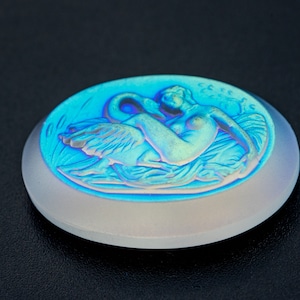 May include: A white oval cameo with an iridescent finish depicting a woman reclining on a swan. The cameo has a raised design and is set against a black background.