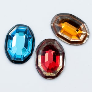 May include: Three colorful, oval-shaped gemstones. One is a vibrant blue, another is a fiery red, and the third is a warm amber color. Each gem has a faceted surface, catching the light and creating a sparkling effect. The gems are set against a white background.