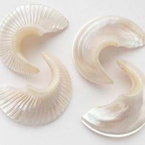 Puede incluir: Tres pendientes de concha de mar de color blanco iridiscente con forma curva y superficie texturizada.