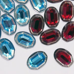 May include: Assortment of oval-shaped, faceted glass rhinestones in shades of blue and red. The rhinestones have a silver-toned backing.