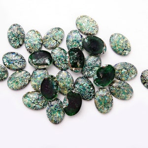 May include: A collection of oval-shaped green and iridescent glass cabochons. The cabochons have a crackled, textured surface and are scattered on a white background.