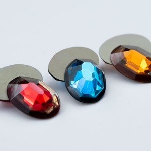 May include: Three oval-shaped rhinestones on gray backing. The rhinestones are red, blue, and orange.
