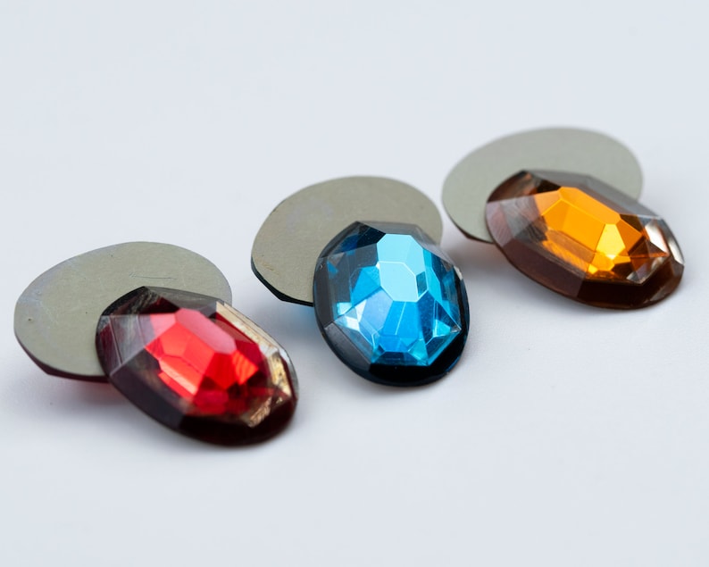 May include: Three oval-shaped rhinestones on gray backing. The rhinestones are red, blue, and orange.