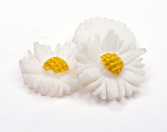 May include: Three white sugar paste daisy flowers with yellow centers. The flowers are arranged in a cluster on a white background.
