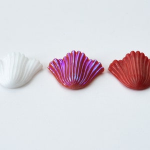 Op de afbeelding: Drie decoratieve schelpvormige versieringen in wit, iriserend roze en rood.