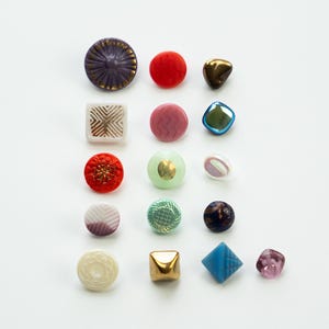 Puede incluir: Una colección de botones y cuentas coloridas de varias formas. El surtido incluye formas redondas, cuadradas y geométricas en tonos morado, rojo, dorado, rosa, verde, azul y blanco. Algunos tienen superficies texturizadas y patrones decorativos.