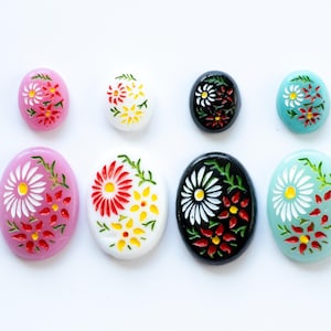 Puede incluir: Surtido de cabujones ovalados con diseños florales. Los cabujones están pintados con flores blancas, rojas, amarillas y negras sobre un fondo rosa, azul, blanco o negro.