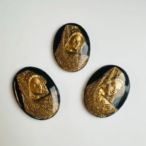 May include: Three oval cameos with a vintage aesthetic. Each has a gold-coloured relief of a woman's profile set against a black backdrop. The woman is wearing an elaborate headdress. The cameos are likely made of resin or a similar material.