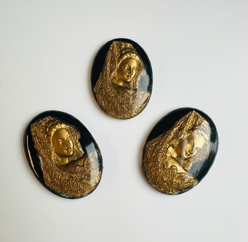 May include: Three oval cameos with a vintage aesthetic. Each has a gold-coloured relief of a woman's profile set against a black backdrop. The woman is wearing an elaborate headdress. The cameos are likely made of resin or a similar material.