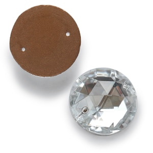 Pode incluir: Um strass redondo de cristal transparente com dois furos para costura. O strass está sobre um suporte marrom.