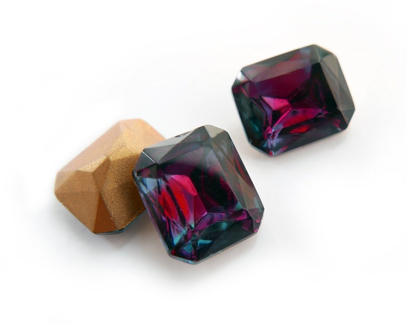 May include: Three faceted gemstones, two are dark purple with a green iridescent sheen and one is gold.