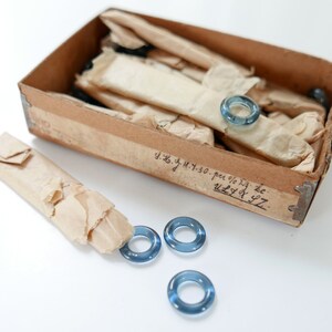 May include: A box of blue glass rings, some are wrapped in brown paper, and some are loose on a white surface. The box is labelled with handwritten text: "y.z.g.y.h. 30. per % 5g. zc. u2.4 x 97."